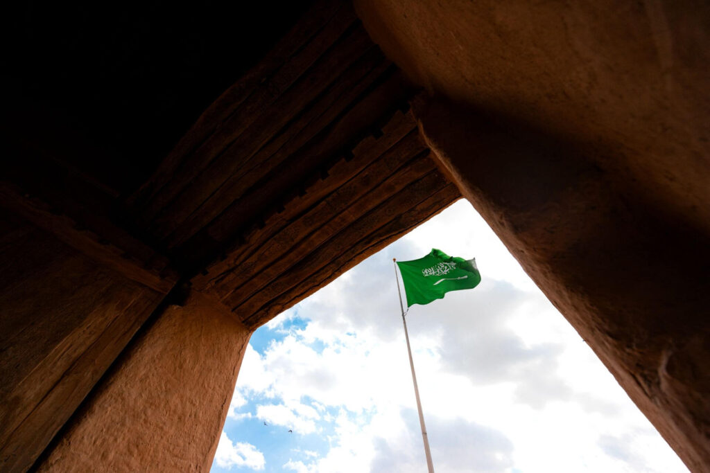 La bandera de Arabia Saudita ondea contra el cielo azul en medio de una arquitectura tradicional.